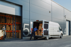 Outside of Logistics Retailer Warehouse With Female Manager Using Tablet Computer, Worker Loading Delivery Truck with Cardboard Boxes. Online Orders, Purchases, E-Commerce Goods, Merchandise